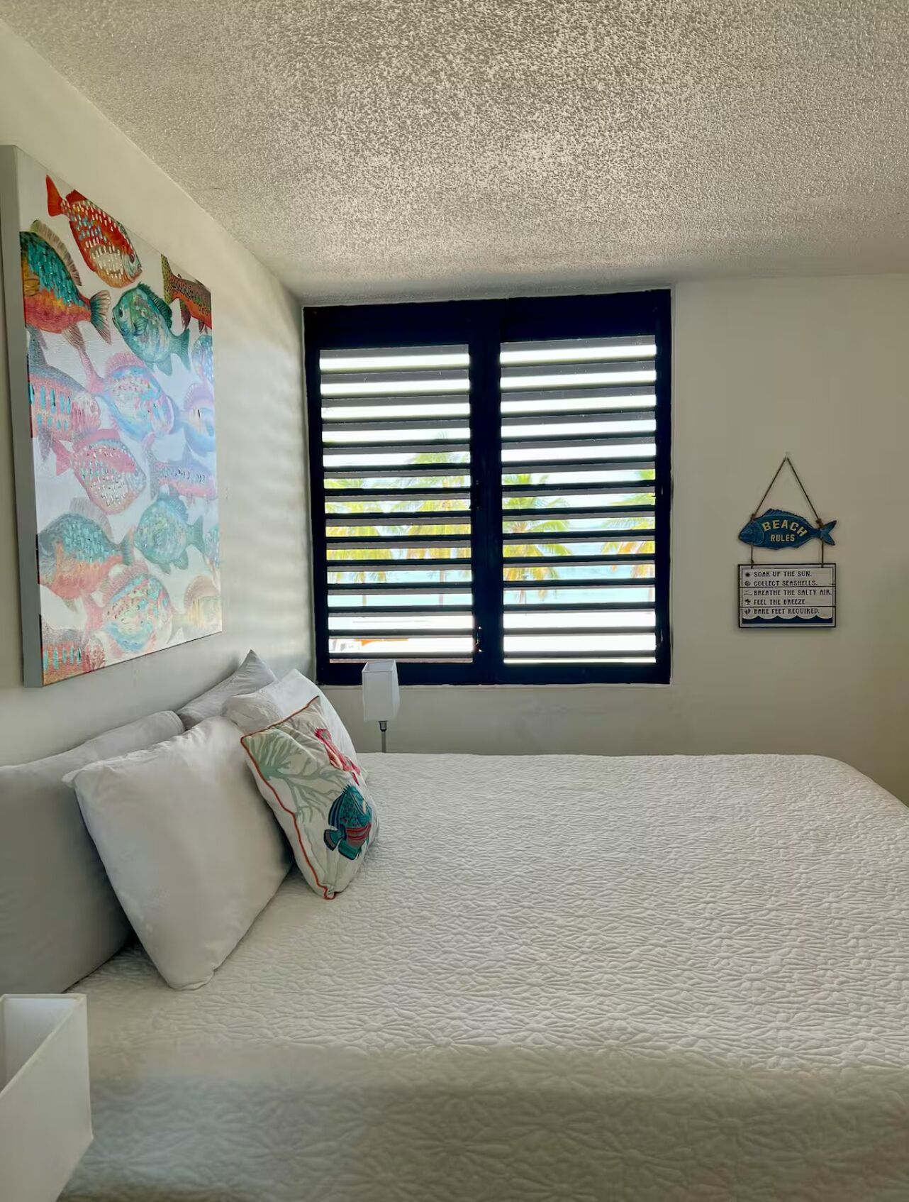 Un dormitorio con una cama grande, una ventana con persianas, una decoración de peces en la pared y un letrero con las normas de la playa.