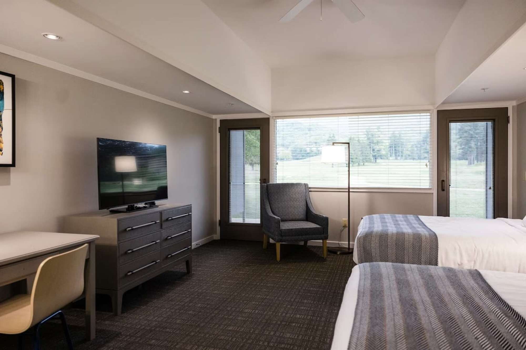 A hotel room with a bed, a desk with a television, a chair, and a window with blinds.