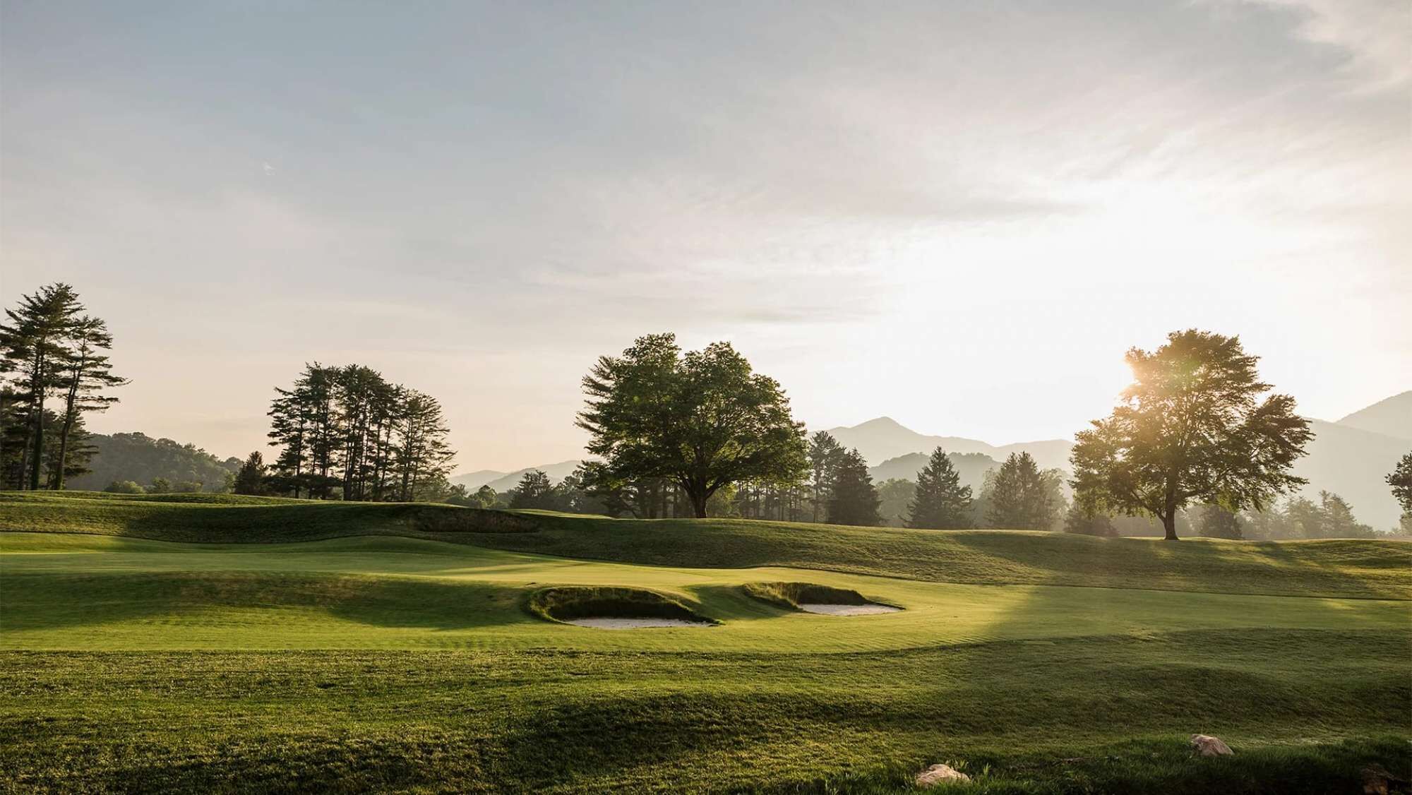 A golf course with rolling hills, trees, and a mountain backdrop.