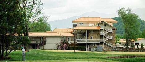 A multi-story building with a stone facade and a balcony, surrounded by a golf course and trees.