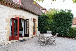 A stone building with a gravel patio, a wooden table and chairs, and a hedge.