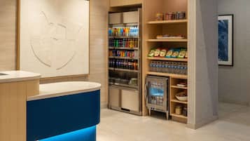 A hotel reception area with a counter, a beverage refrigerator stocked with drinks, and shelves with snacks.
