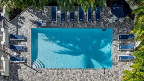 Una piscina rectangular amb una terrassa enrajolada, rodejada de gandules i ombrel·les, i palmeres al fons.