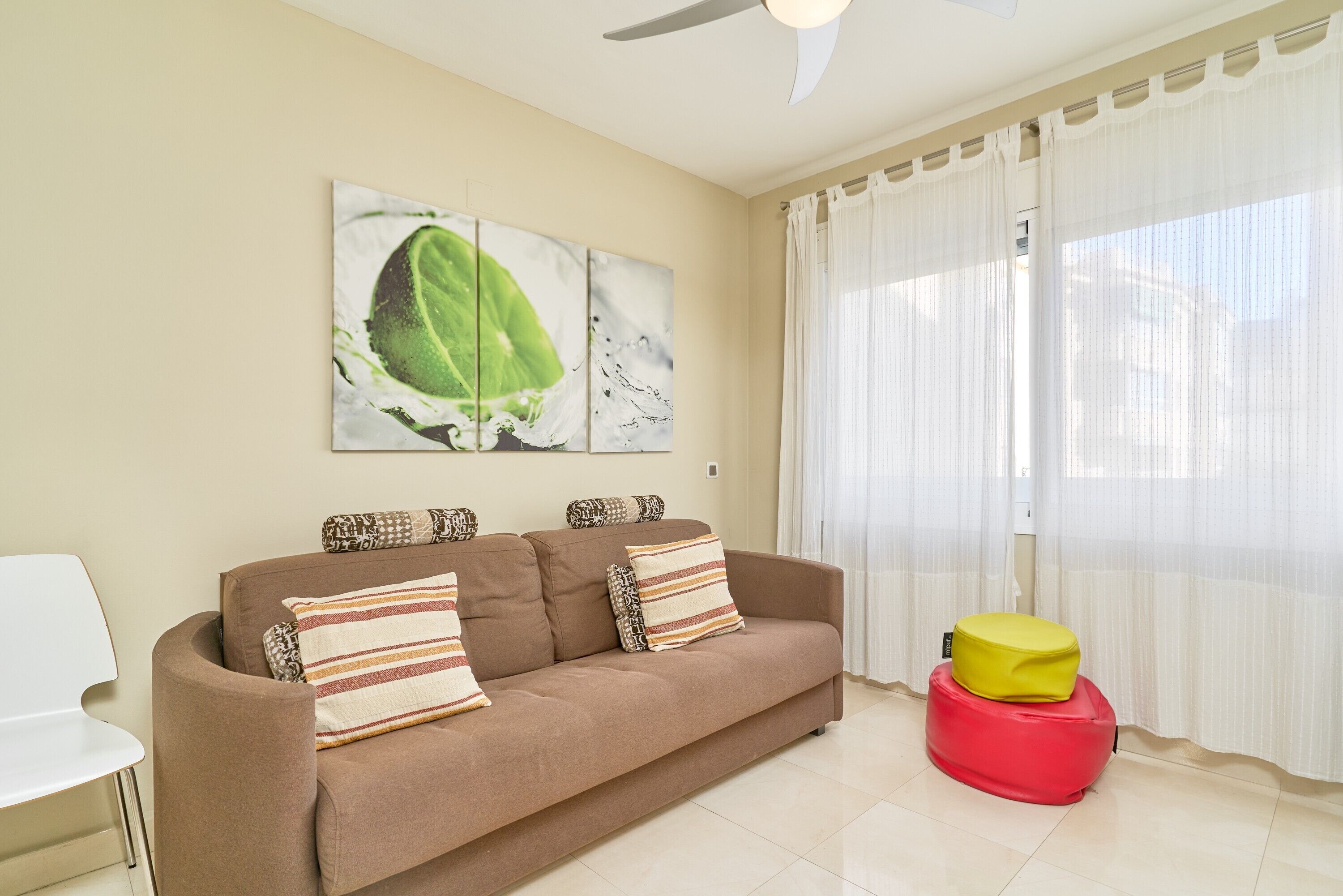A living room with a sofa, pillows, a red ottoman, and a ceiling fan.
