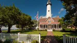 A lighthouse with a brick base and white walls, surrounded by a white picket fence and lush greenery. - Direct Golf Course Access + Pool. Dining. Shuttle. (St. Simons Island)