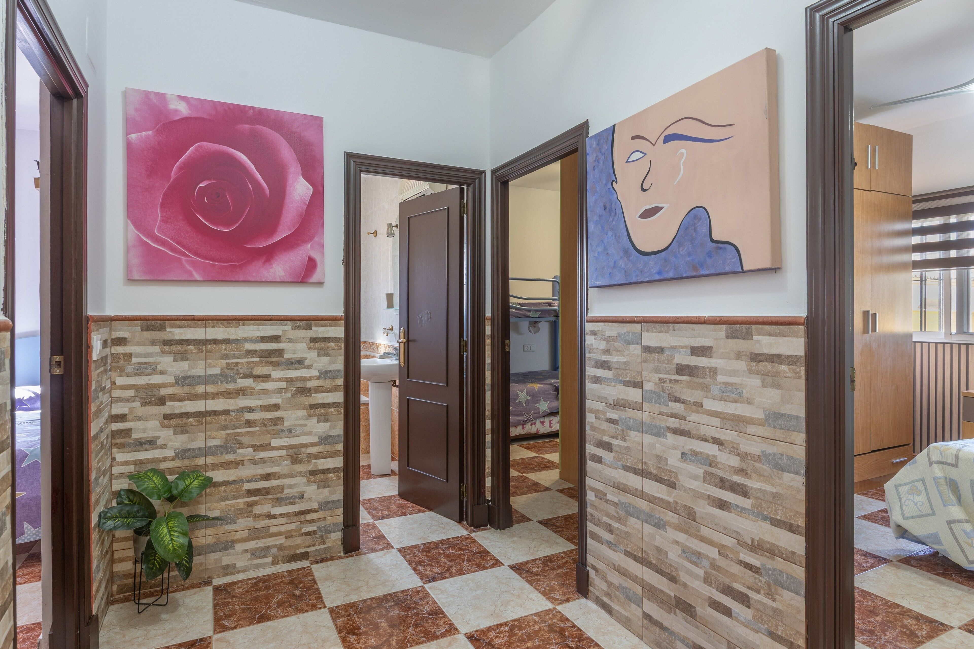 Un pasillo con piso a cuadros, una planta en maceta, una puerta que da al baño y un cuadro en la pared.