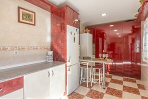 A kitchen with a refrigerator, coffee maker, two bar stools, and a tiled floor.