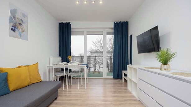 A modern living room with a grey sofa, a dining table with chairs, a flat-screen TV, and a large window with curtains. - Bright Apartment with Bathtub | Balcony and Good Communication (Warszawa)
