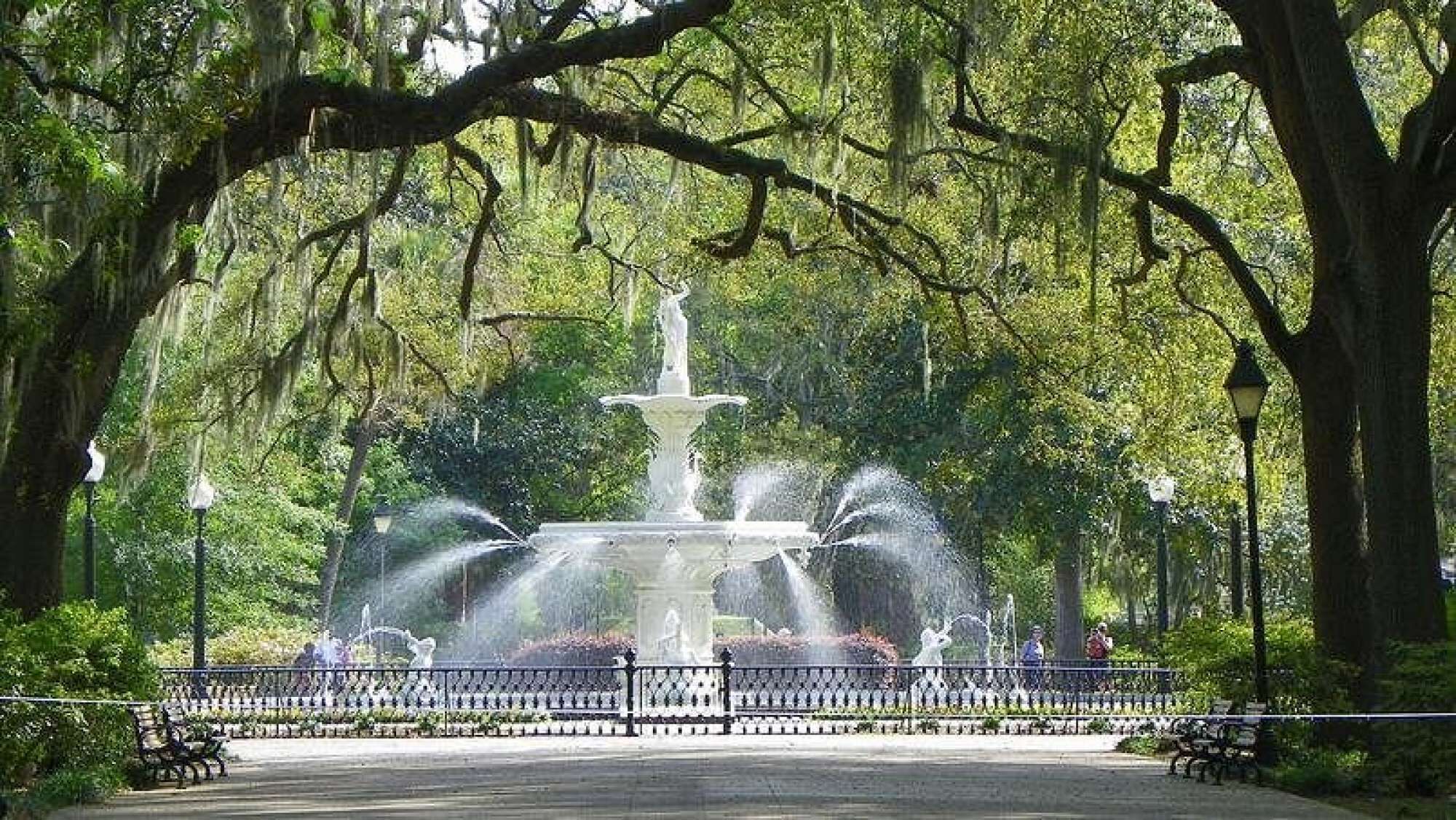 A park with a central fountain, surrounded by benches and trees.