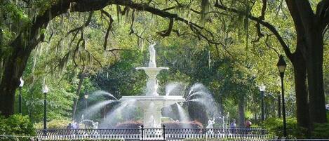 A park with a central fountain, surrounded by benches and trees.