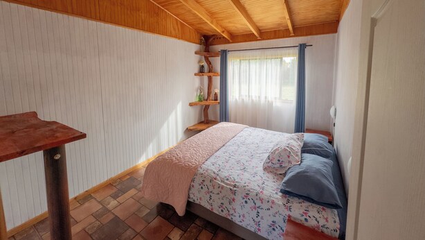 Une chambre avec un plafond en bois, un lit recouvert d’une literie à motifs floraux, une table de chevet et une fenêtre avec des rideaux.