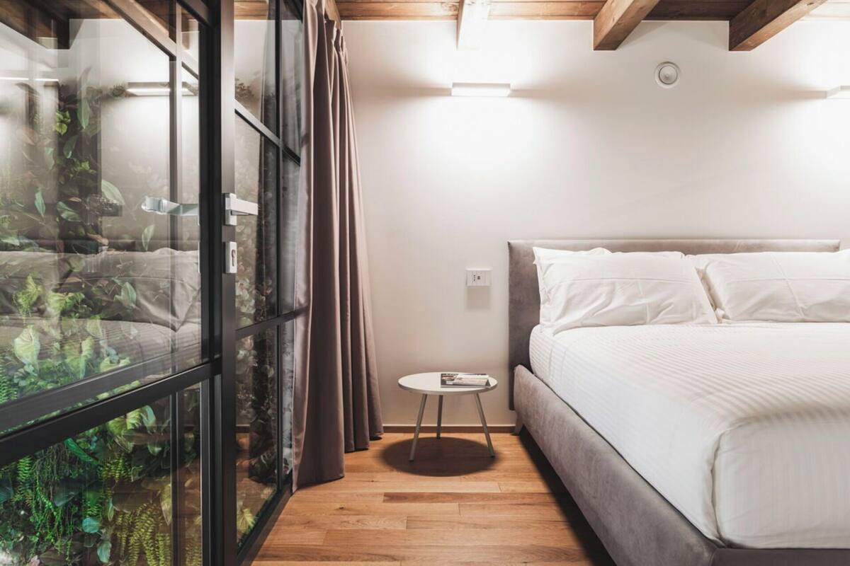 A bedroom with a bed, a small round table, a glass wall with plants, and a ceiling light.