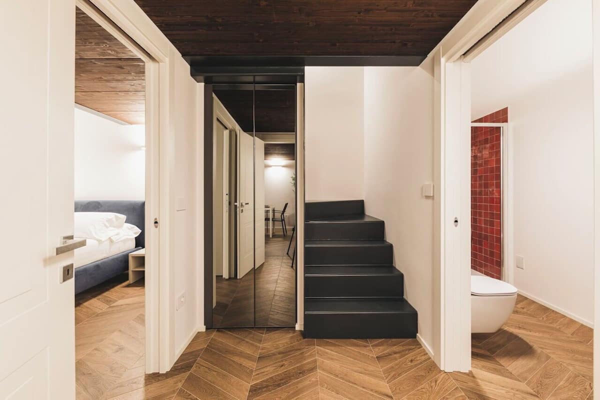 A hallway with a wooden floor, a staircase, and a white door leading to a bathroom with red tiles.