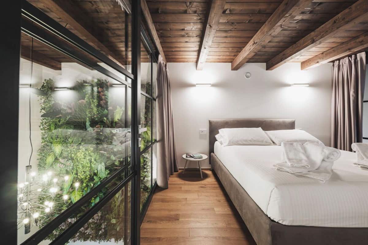 A bedroom with a bed, bedside table, and a view of a garden through a glass wall.