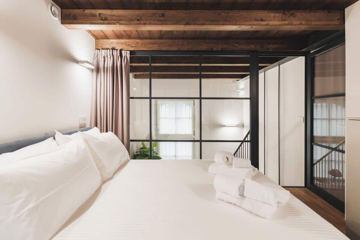 A modern hotel room with a large bed, a wooden ceiling, and a view of the city through floor-to-ceiling windows.