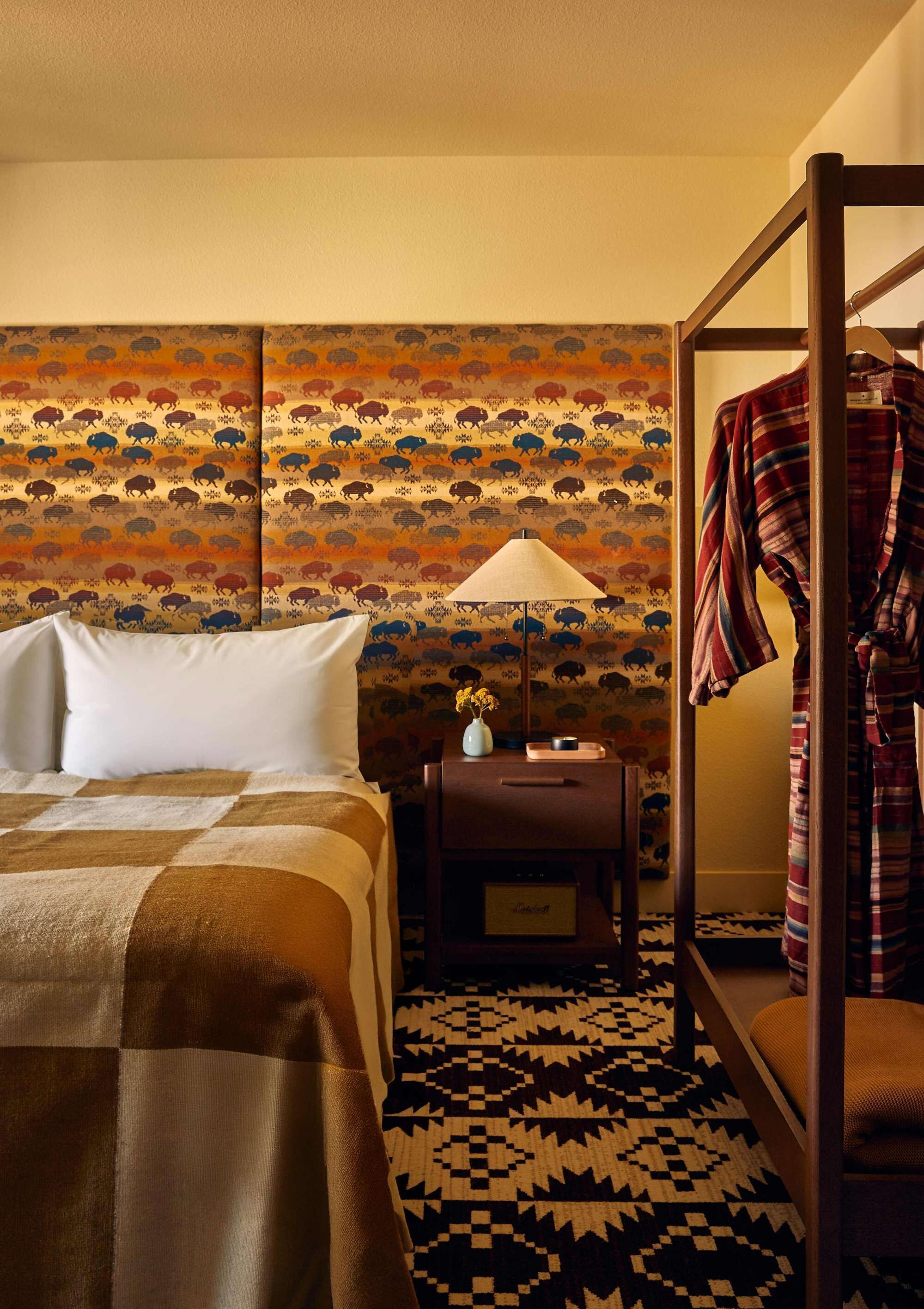 Une chambre d’hôtel avec un lit, une table de chevet, une lampe, un mur à motifs, un tapis à motifs et une robe suspendue.