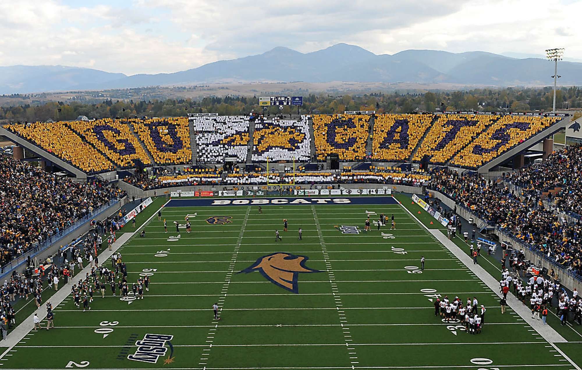 A football stadium with a large crowd, a visible scoreboard, and a mountain backdrop.