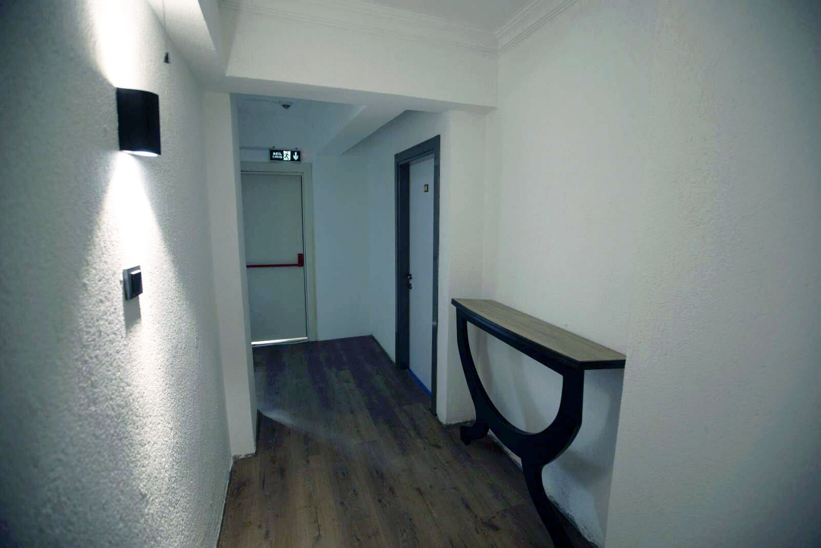 Een smalle gang met een houten vloer, een wandlamp en een deur aan het einde.