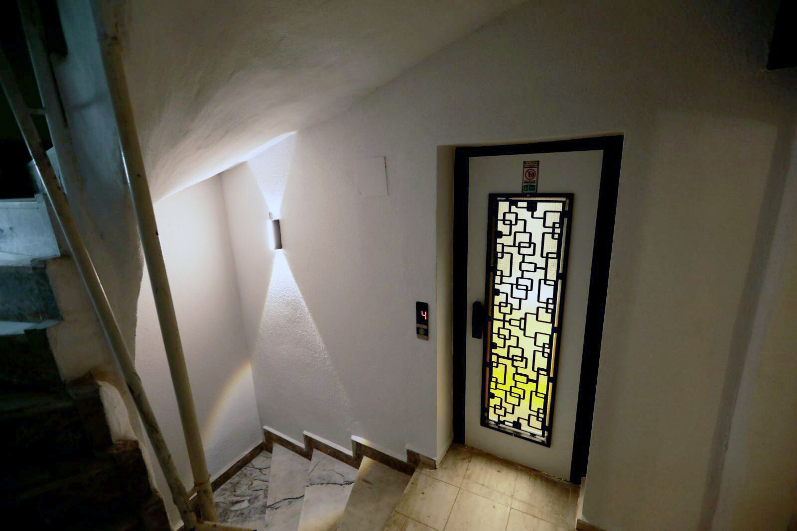 Un pasillo con una escalera a la izquierda, una lámpara empotrada en la pared iluminada, una puerta con un diseño geométrico y un reloj digital que muestra la hora.