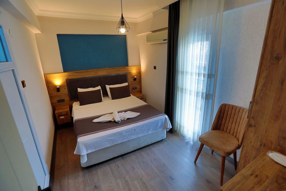 Une chambre d’hôtel comprenant un lit, une chaise, une tête de lit en bois, un climatiseur fixé au mur et une fenêtre avec des rideaux.
