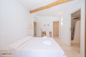 Habitació d'hotel amb un llit, una biga de sostre de fusta, una taula petita amb tovalloles, zona de dutxa i una porta que condueix a una altra habitació.