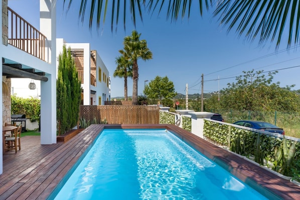 A swimming pool with a wooden deck, surrounded by white buildings, palm trees, and a clear sky.