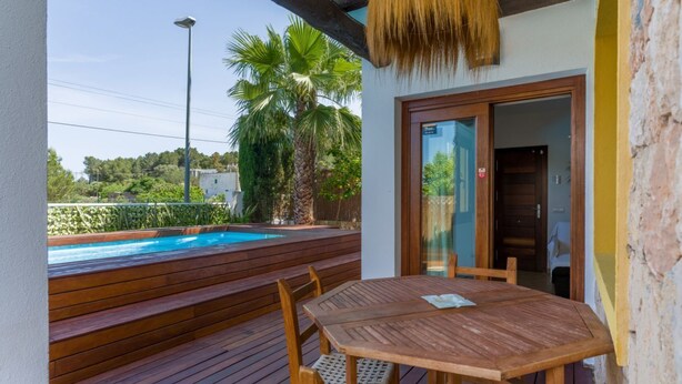 Una terraza de madera con piscina, una mesa con sillas y una puerta que da a un espacio interior.