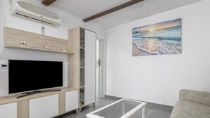 Ein Wohnzimmer mit Flachbildfernseher, einem Glastisch, einem beigen Sofa, einem Holzschrank und einem Gemälde, das einen Strand bei Sonnenuntergang zeigt.