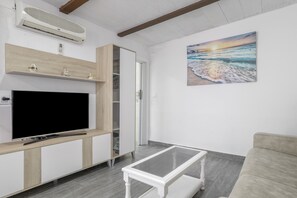 Ein Wohnzimmer mit Flachbildfernseher, einem Glastisch, einem beigen Sofa, einem Holzschrank und einem Gemälde, das einen Strand bei Sonnenuntergang zeigt.