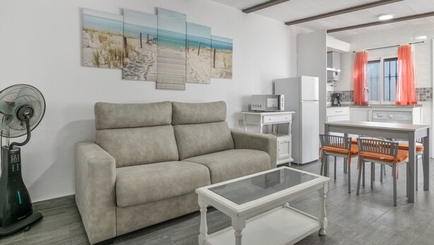 Ein Wohnzimmer mit einer Couch, einem Glastisch, einem Ventilator und einer Küchenzeile mit Esstisch und Stühlen.