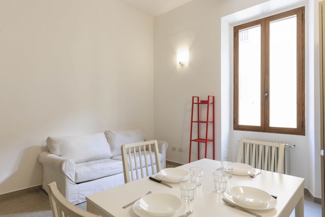 A dining area with a white table set for four, a red chair, and a white sofa in the background.