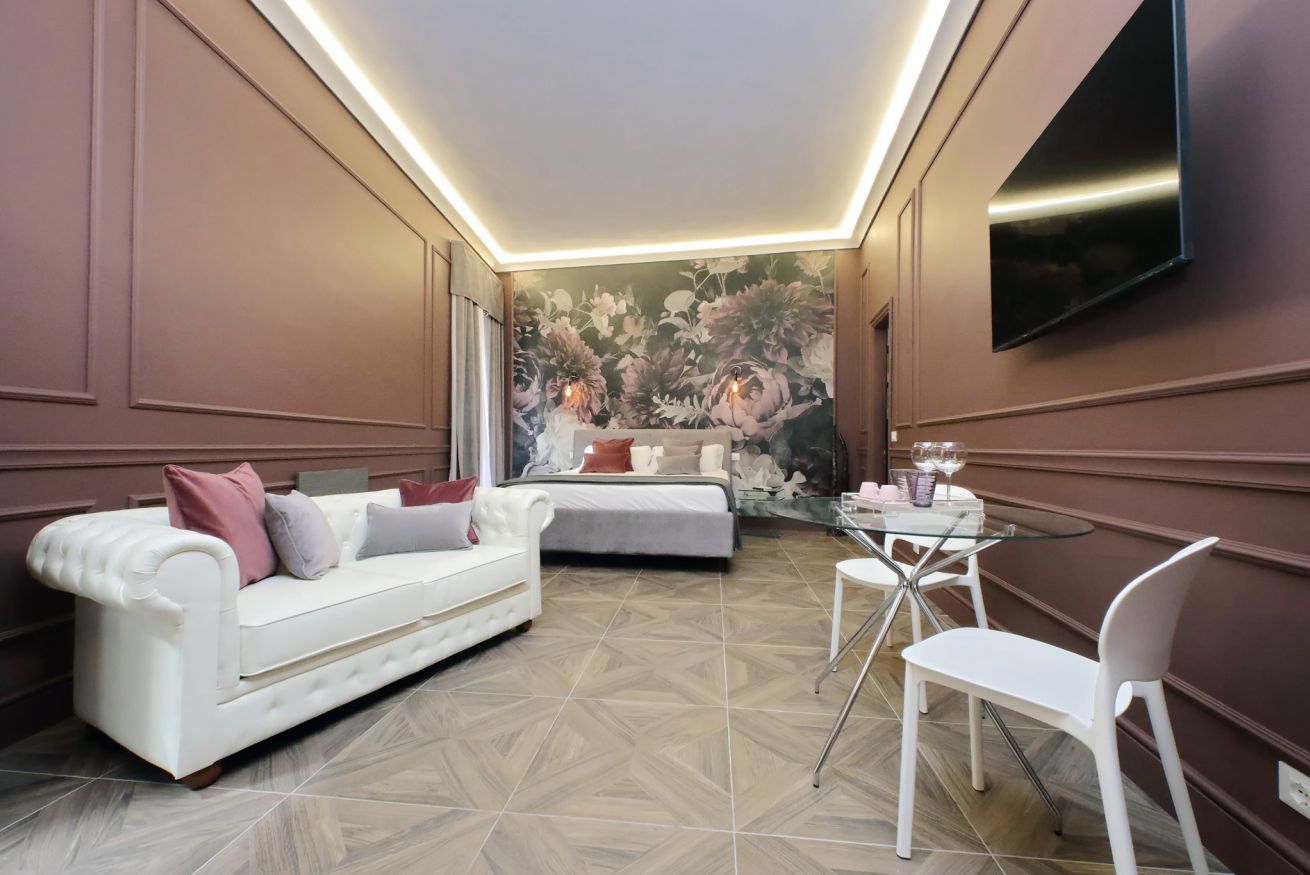 Una habitación de hotel moderna con un sofá blanco, una mesa de comedor de cristal y un televisor grande en la pared.