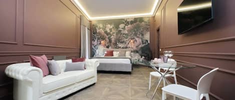 Una habitación de hotel moderna con un sofá blanco, una mesa de comedor de cristal y un televisor grande en la pared.