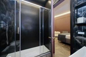 Quarto de hotel moderno com chuveiro em cabine de vidro, cama com colcha cinza e estante com objetos decorativos.