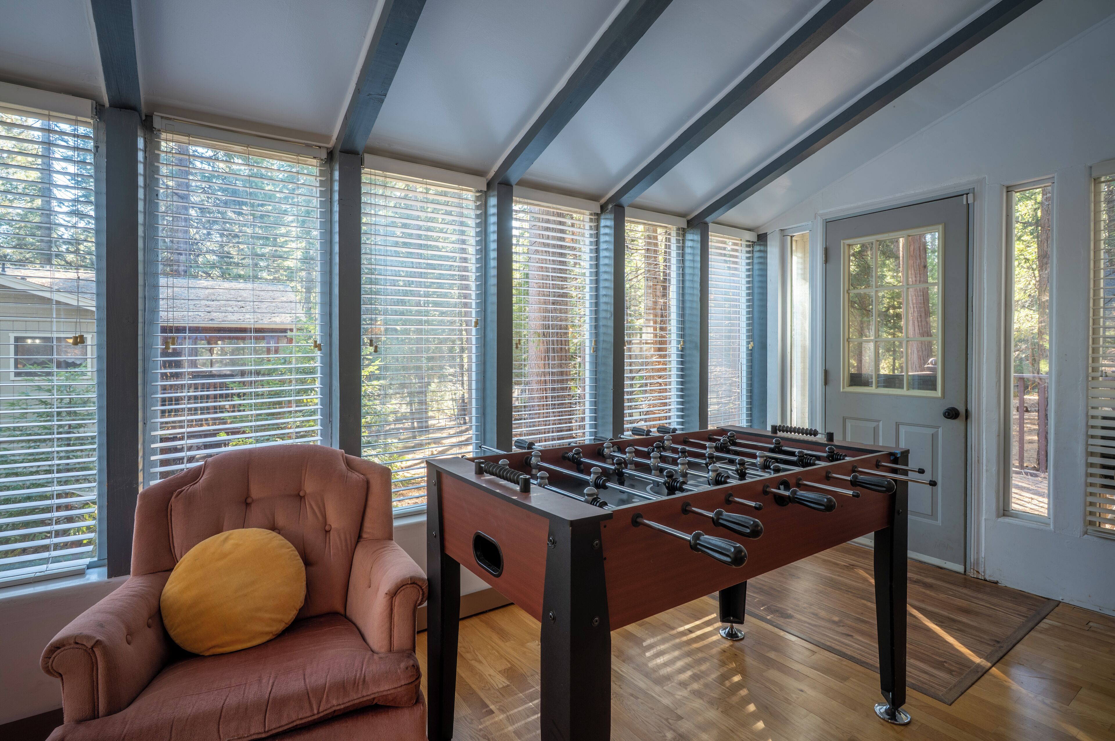 A room with a foosball table, a leather armchair, and large windows with blinds.