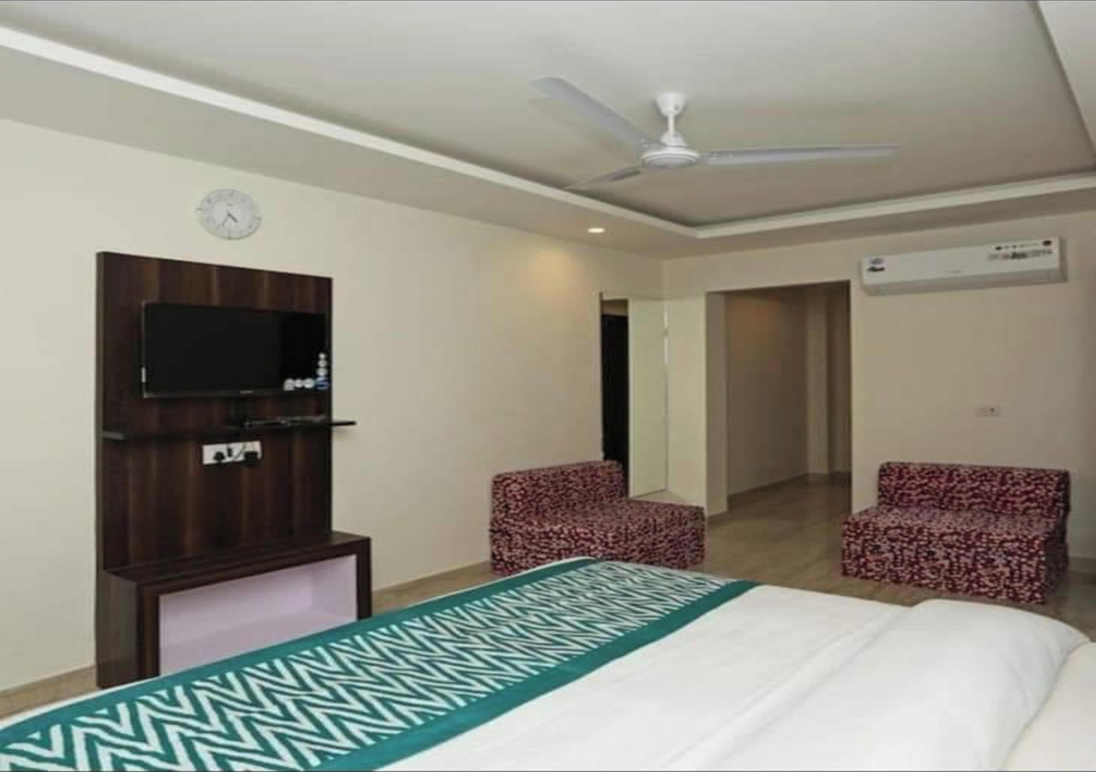 A hotel room with a bed, a TV mounted on the wall, two patterned chairs, a ceiling fan, and an air conditioning unit.