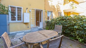 A patio with a wooden table and chairs, a yellow building with a door, and a hedge. - Rental in Rome Pateras Garden Apartment (Rome)