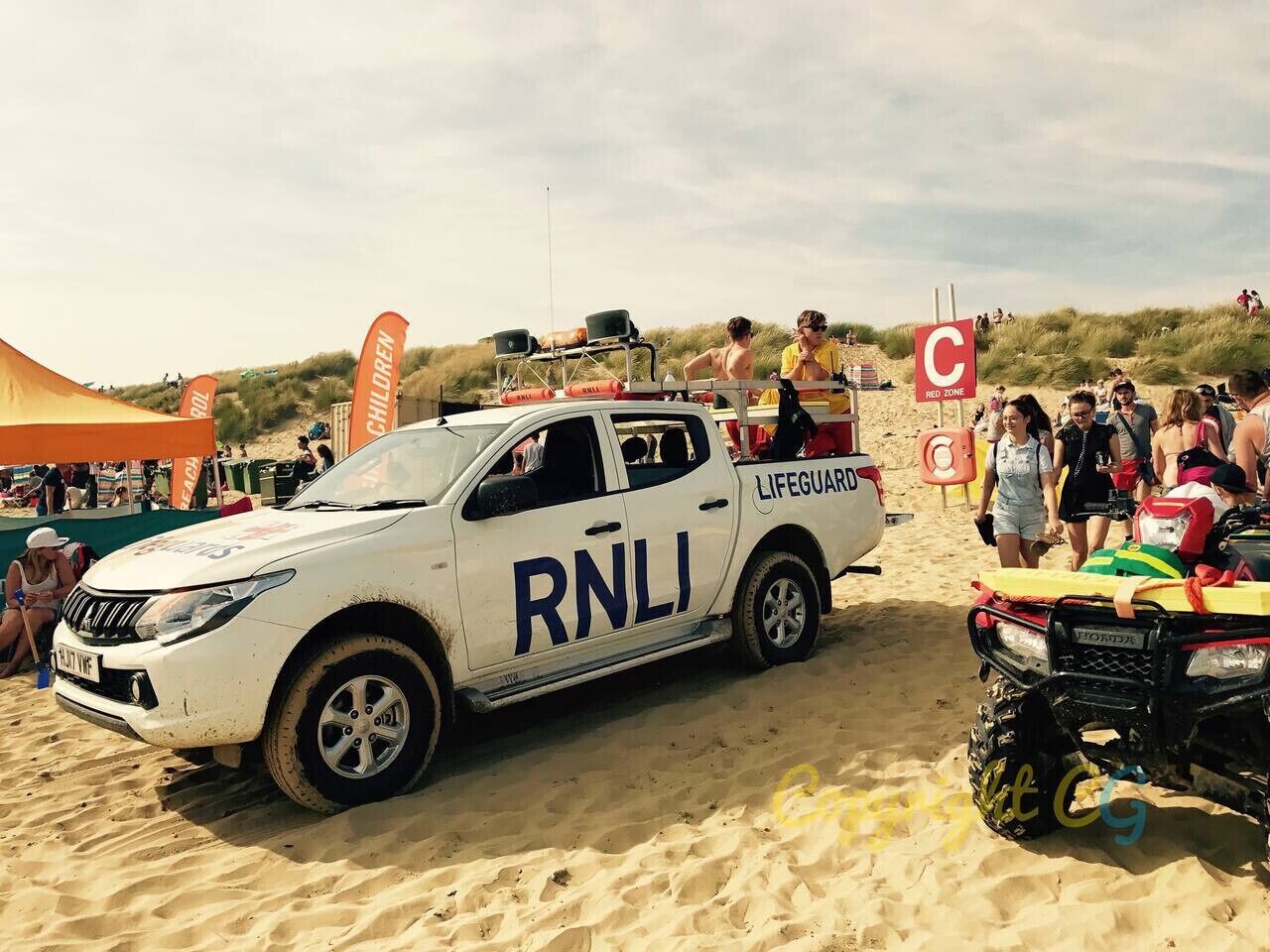 Un vehículo de socorristas blanco con las leyendas "RNLI" y "LIFEGUARD" en la carrocería, estacionado en una playa arenosa con gente y un cartel de zona roja al fondo.