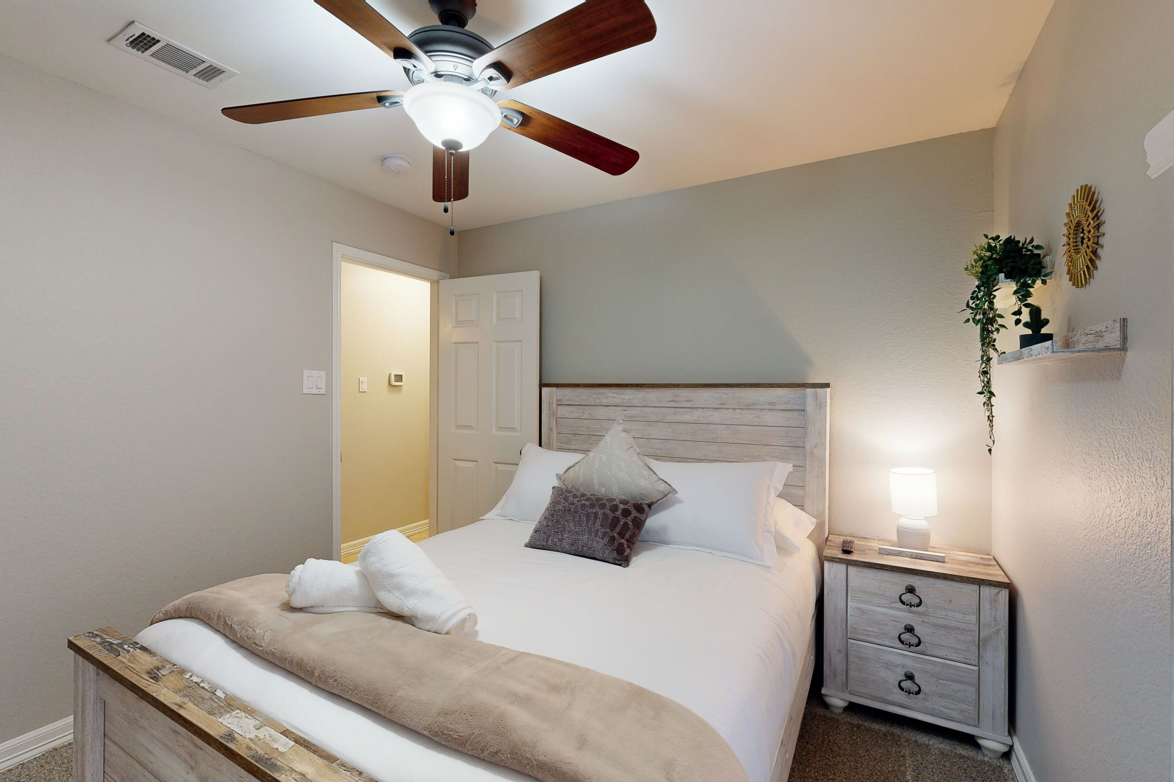 Un dormitorio con cama, ventilador de techo, mesita de noche con lámpara y puerta que da a otra habitación.