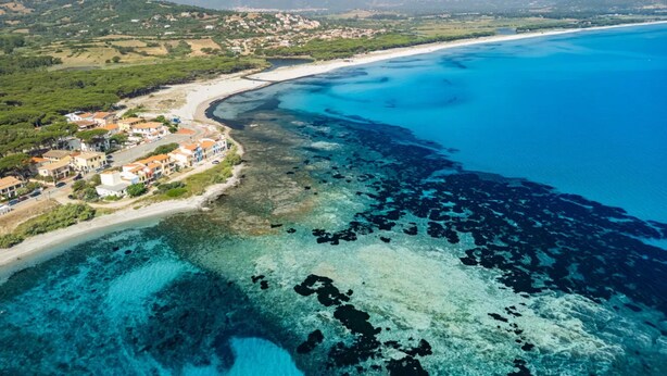 Vista aerea di una cittadina costiera con acque cristalline, una spiaggia sabbiosa e edifici con tetti rossi.