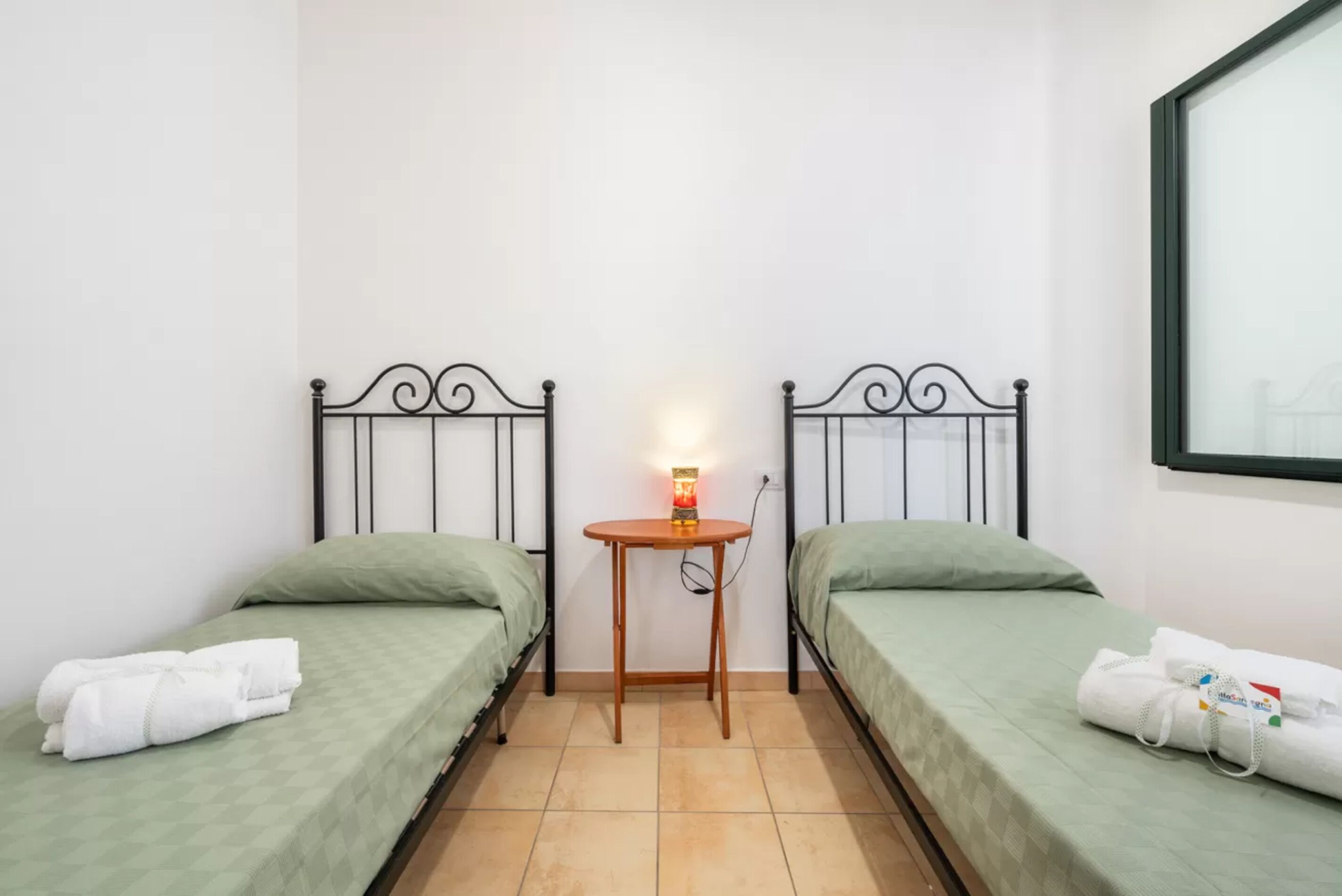 Ein kleines, sauberes Zimmer mit zwei Einzelbetten, einem kleinen Holztisch mit Lampe und einem Spiegel an der Wand.