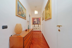 A hallway with wooden flooring, a wooden dresser with a woven lamp, and framed paintings on the walls. - UNA FINESTRA SULLA CUPOLA DI SAN PIETRO (Roma)