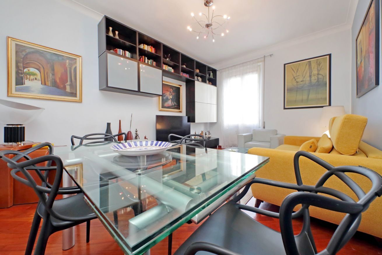 A modern living room with a glass dining table, black chairs, a yellow sofa, and a bookshelf.