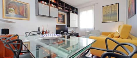 A modern living room with a glass dining table, black chairs, a yellow sofa, and a bookshelf.