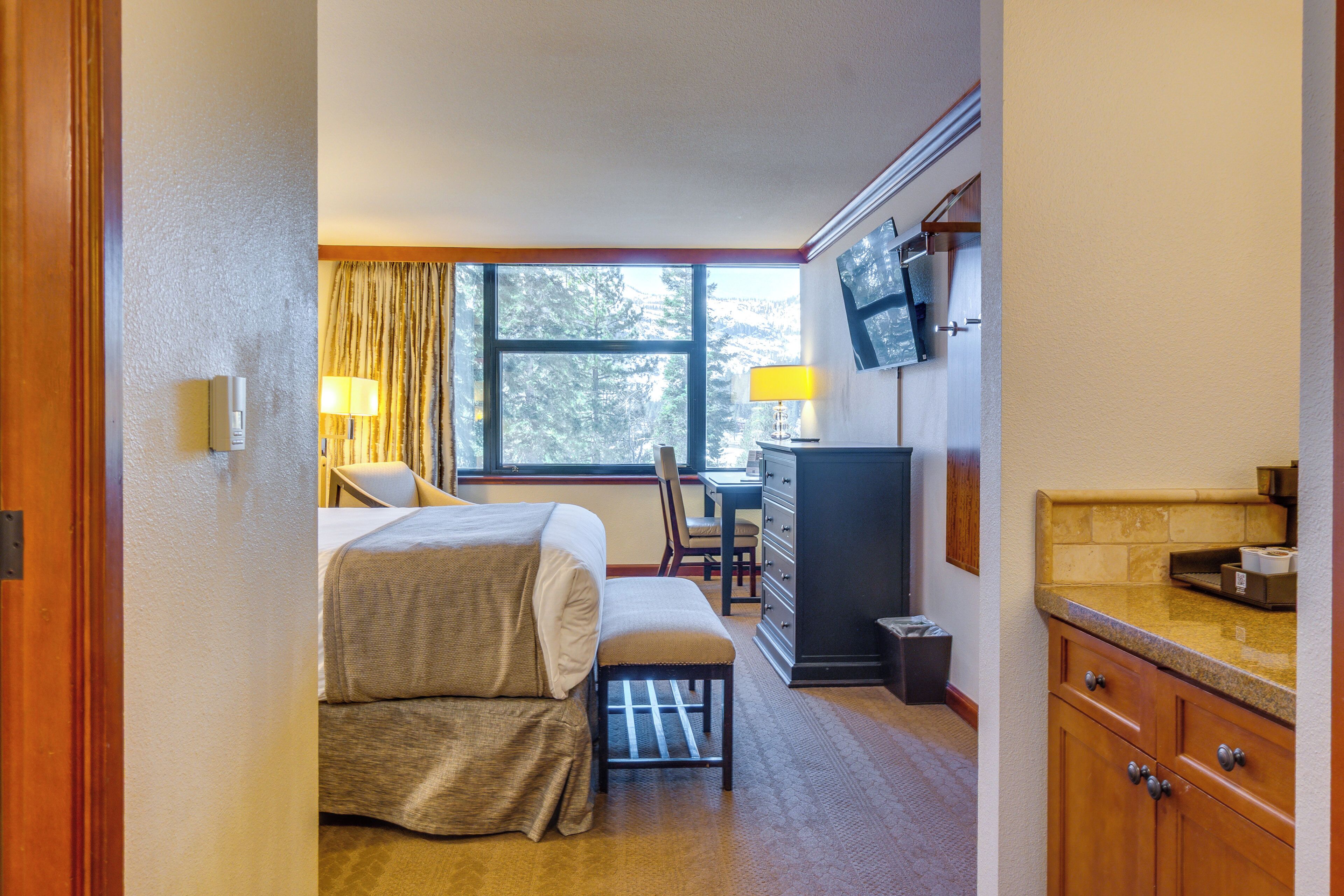 A hotel room with a bed, a desk, a chair, a TV, and a kitchen area.