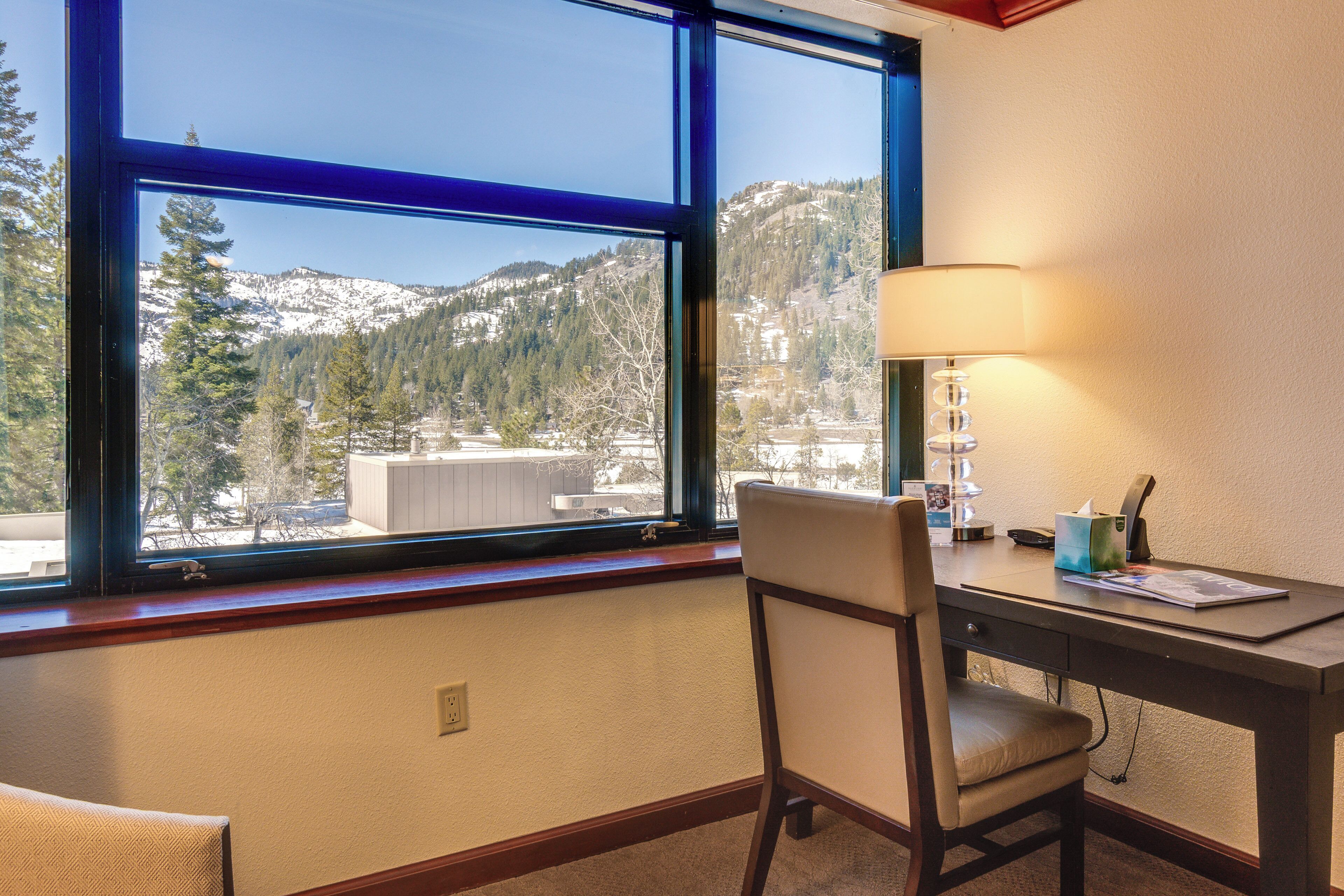 A room with a desk, chair, and a large window offering a view of snow-covered mountains and trees.