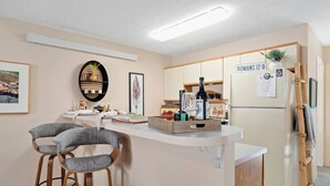 A kitchen with a white refrigerator, wooden bar stools, a white countertop, and a wall-mounted shelf with decorative items. - Walk to Midtown Ruidoso! Cozy Eagles Nest Condo (Ruidoso)