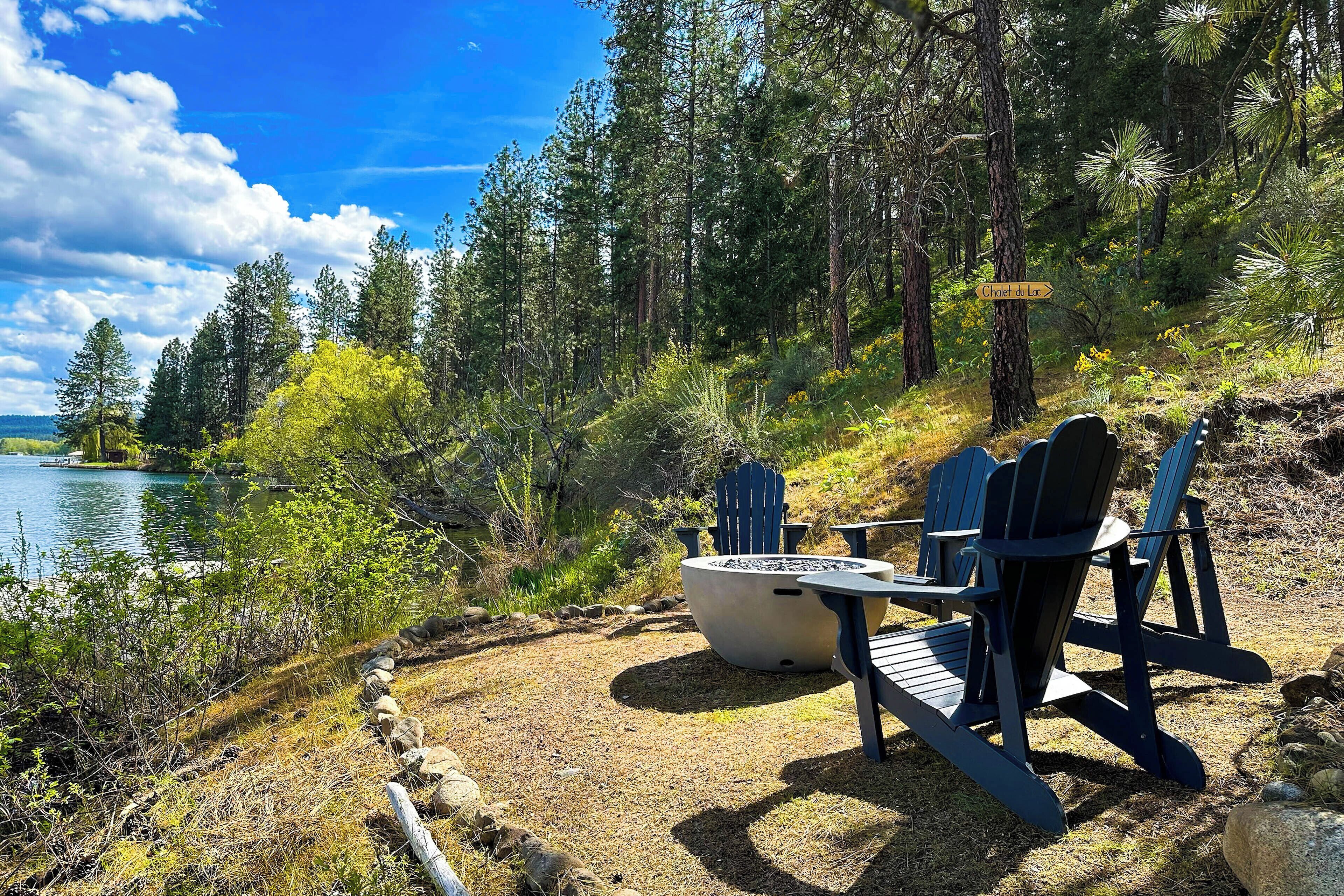 Ein Sitzbereich am See im Freien mit Adirondack-Stühlen und einem Feuerkorb, umgeben von einer bewaldeten Landschaft.