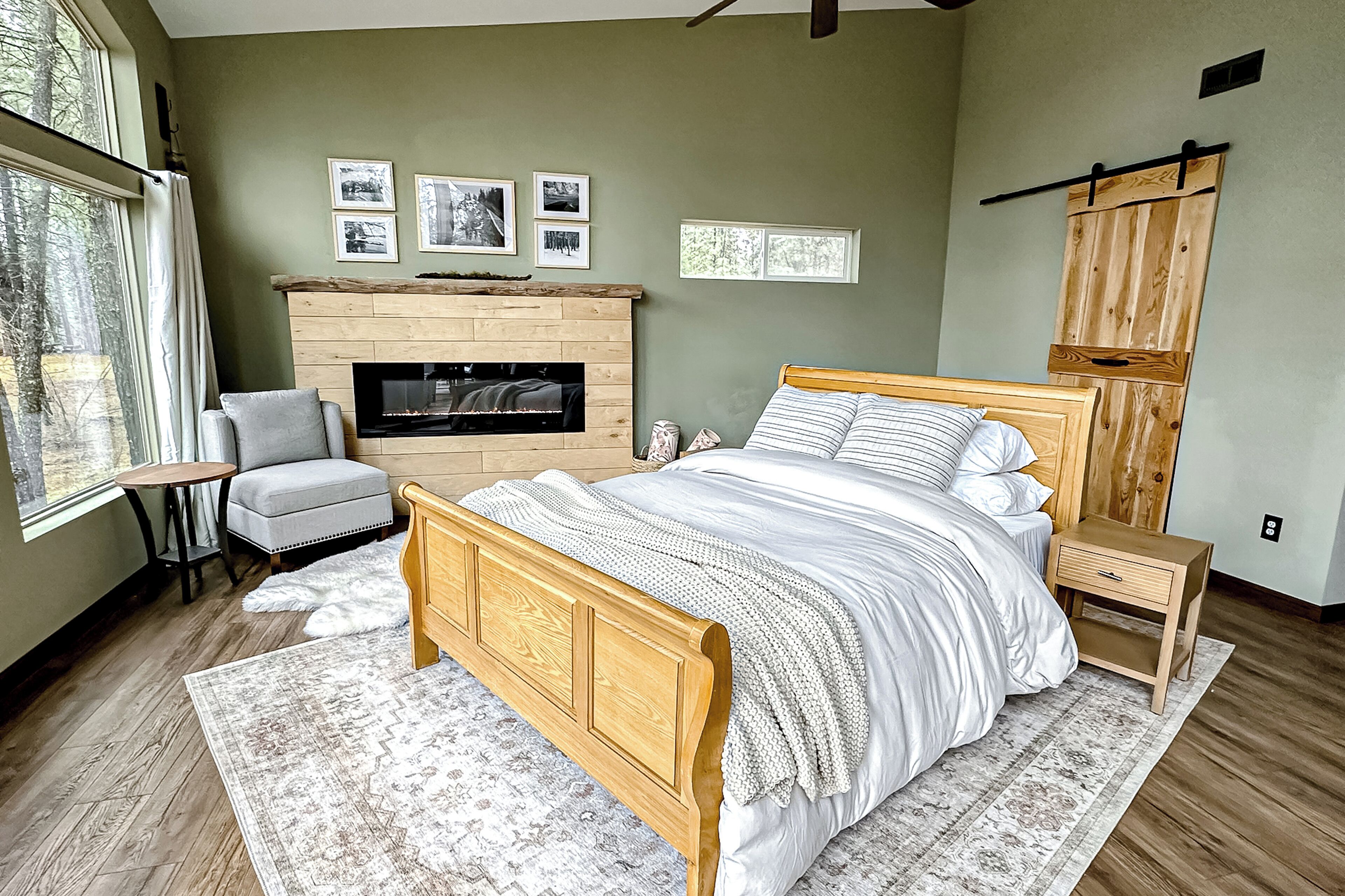 A bedroom with a wooden bed, a fireplace, a chair, and a view of trees through a window.