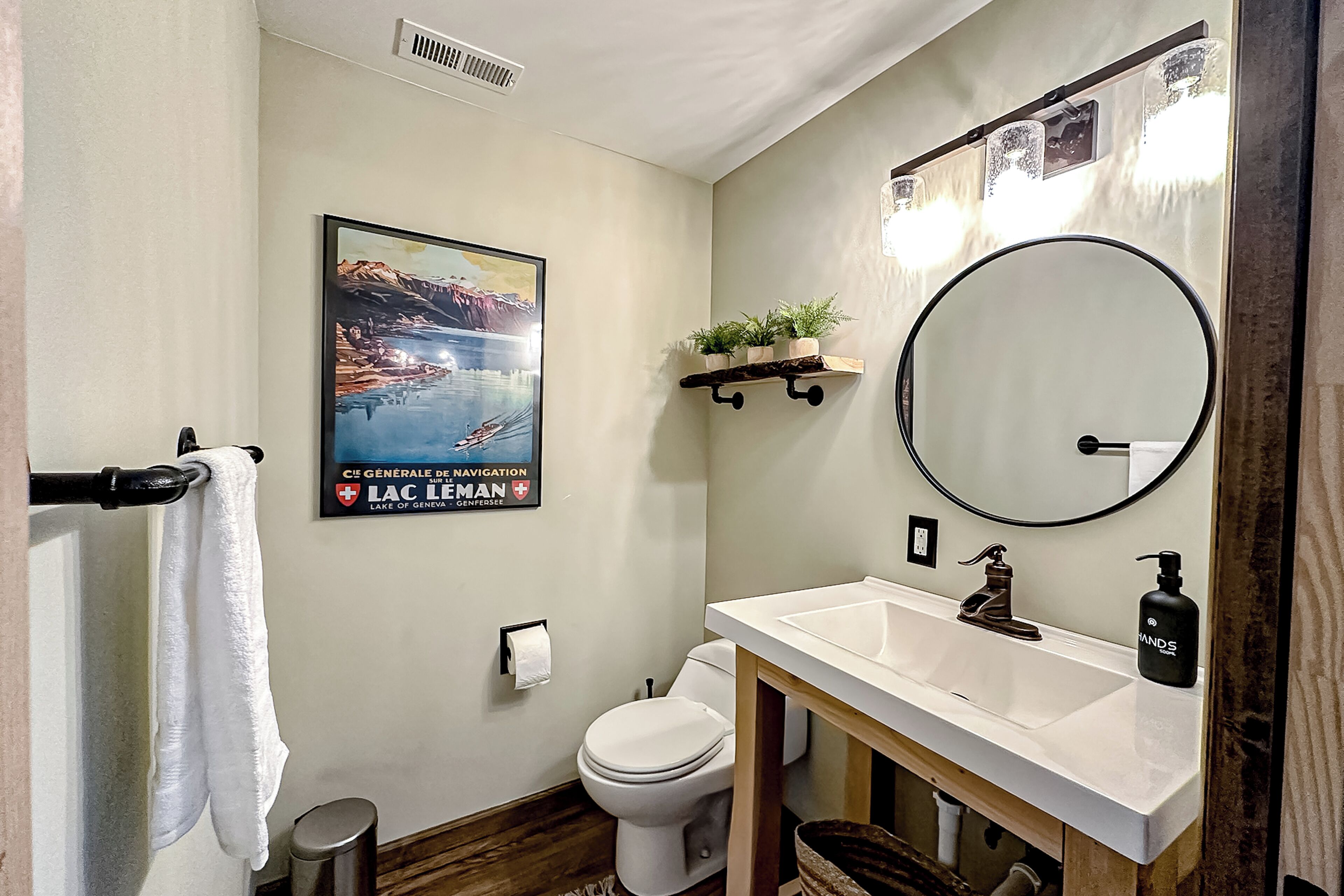 Ein Badezimmer mit weißem Waschbecken, einem runden Spiegel, einer Toilette, einem Handtuchhalter, einem Mülleimer und einem gerahmten Bild an der Wand.